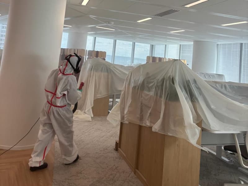 Personne en combinaison et masque, pulvérisateur en main, entre des bureaux recouverts de bâches plastiques