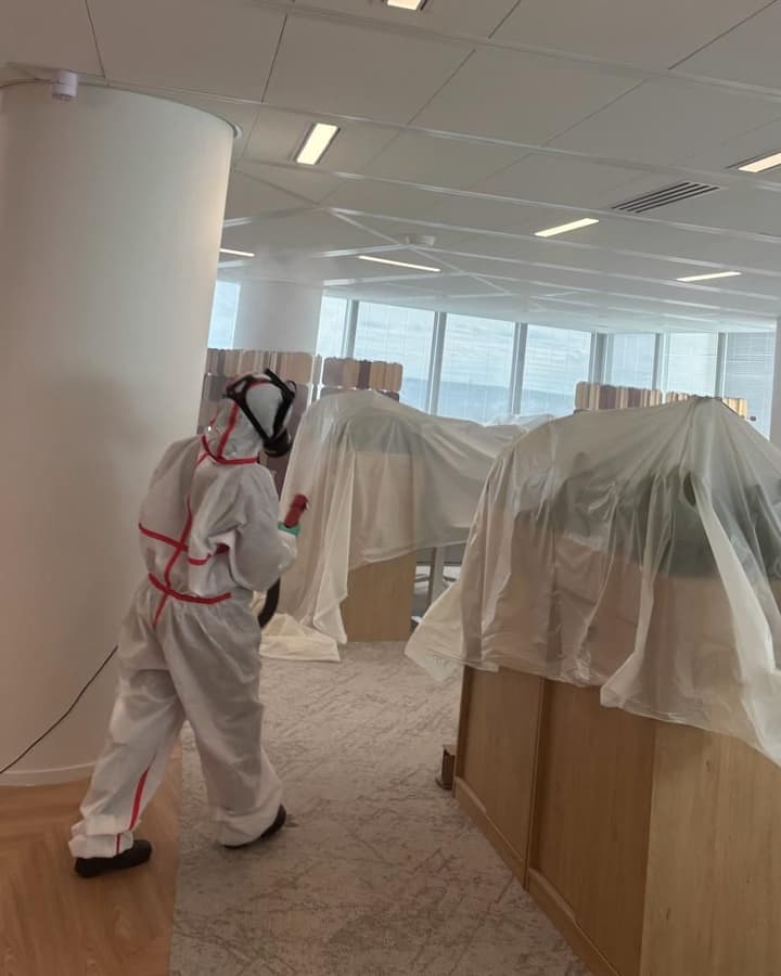 Personne en combinaison et masque, pulvérisateur en main, entre des bureaux recouverts de bâches plastiques