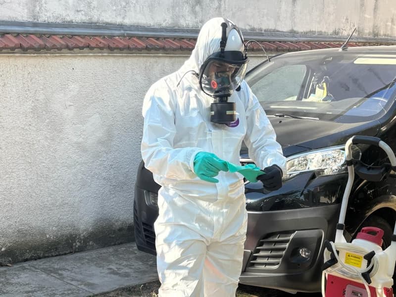 Personne en combinaison blanche et masque intégral, gants verts en main, près d'un pulvérisateur sur roues et d'un véhicule