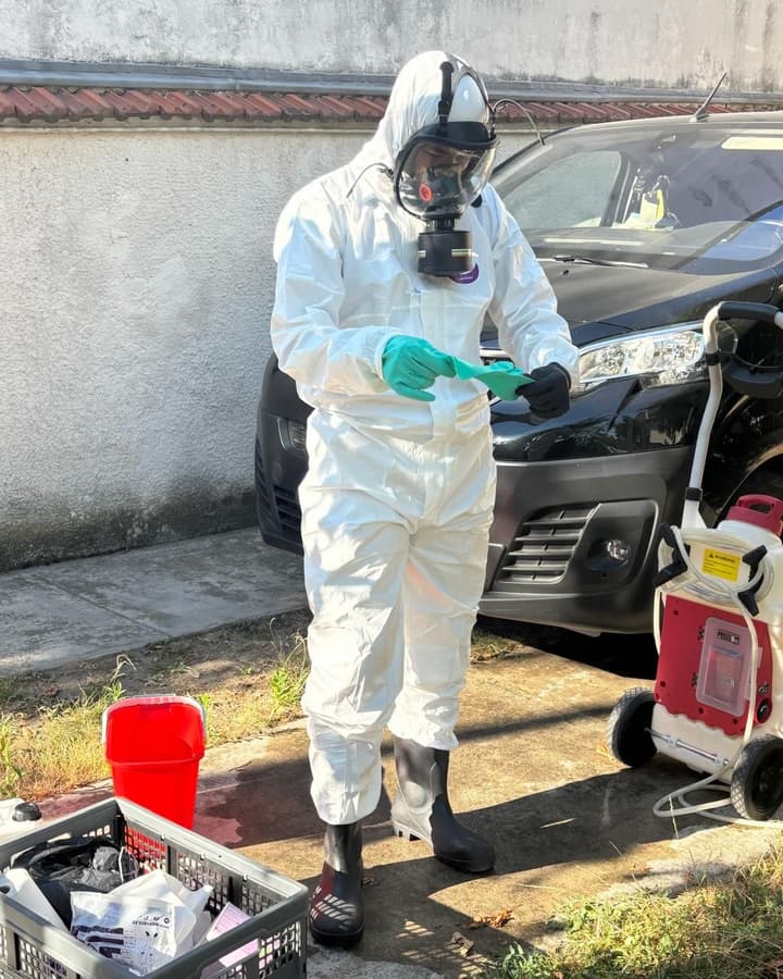 Personne en combinaison blanche et masque intégral, gants verts en main, près d'un pulvérisateur sur roues et d'un véhicule