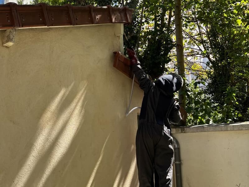 Personne en combinaison sombre à capuche intégrale, bras levés, dirigeant une lance entre les tuiles et le mur